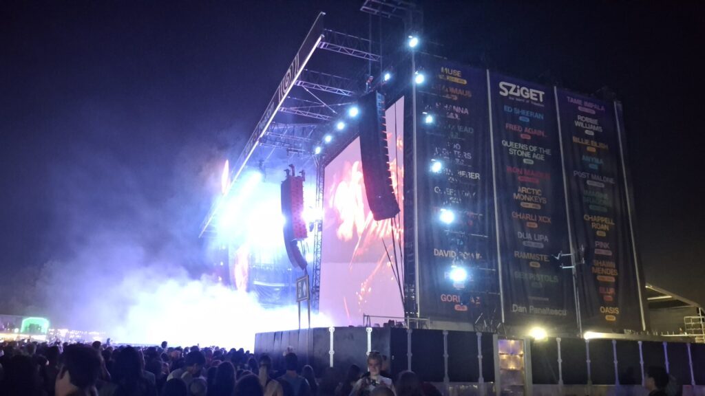 Nachtopname van een groot festivalpodium tijdens het Sziget Festival. Schijnwerpers en ledschermen verlichten het podium, terwijl zich ervoor een dichte menigte verzamelt. Aan de zijkant van de podiumconstructie hangen grote banners met het Sziget-logo en de namen van verschillende artiesten.